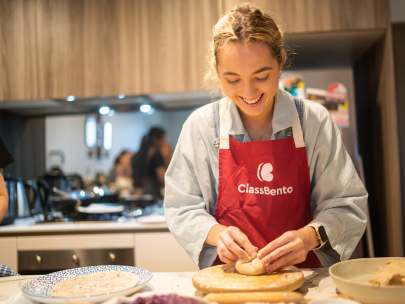 A woman in a red ClassBento apron is making a flatbread at an Indian cooking class
